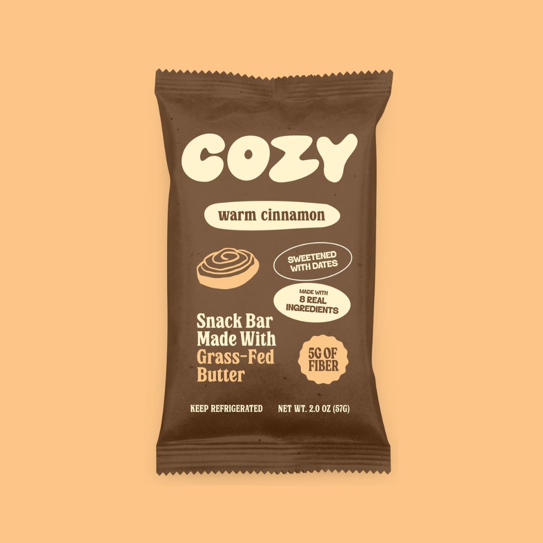 A single Cozy brand 'warm cinnamon' snack bar wrapper, made with grass-fed butter and 5g of fiber, shown on a solid peach background.