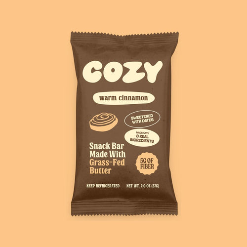 A single Cozy brand 'warm cinnamon' snack bar wrapper, made with grass-fed butter and 5g of fiber, shown on a solid peach background.
